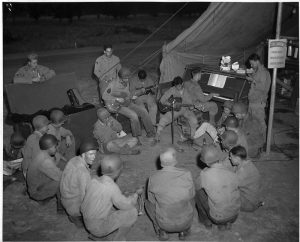 Soldiers in the field in WW2 gathered around two soldiers, one playing a guitar and the other playing a GI Steinway piano.